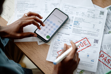 Black young man analyzing invoices and bills using smartphone calculator and pen, hands holding pen...