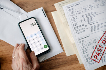 Adult man using smartphone calculator while analyzing overdue utility bills and invoices on wooden desk, hand interacting with device and paperwork