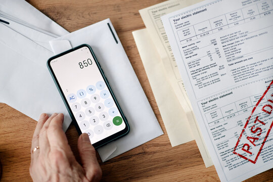 Adult man using smartphone calculator while analyzing overdue utility bills and invoices on wooden desk, hand interacting with device and paperwork