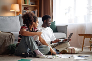 Young Black man using laptop while young Black woman analyzing invoices and bills on floor in living room, both sitting cross legged surrounded by paperwork and documents