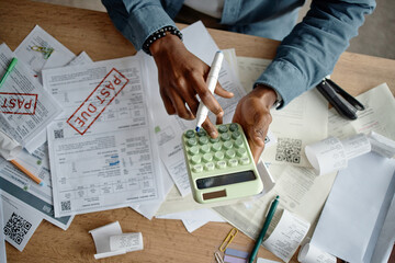 Black man using calculator and pen while analyzing invoices and bills on cluttered desk, reviewing...