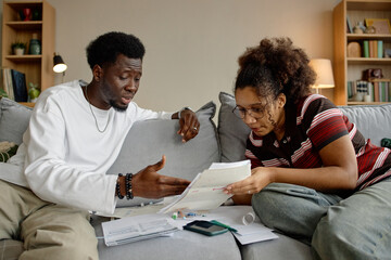 Young Black man and young Black woman sitting on sofa analyzing invoices and bills together, discussing financial documents with calculator and paperwork spread on table in living room