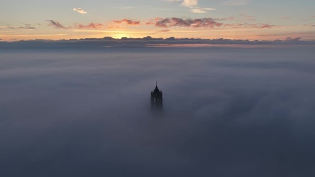 Cinematic aerial video of church tower rising from low passing clouds and fog during sunrise in the city of Utrecht, the Netherlands