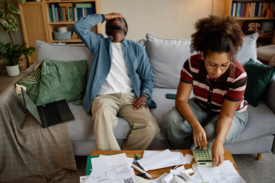 Young Black man sitting on couch holding head in frustration while couple analyzing invoices and bills at home - Powered by Adobe