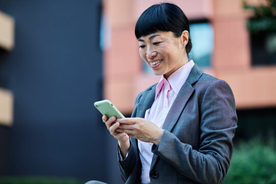 Portrait of a young asian japanese businesswoman woman using a smartphone mobile phone walking down the street, surrounded by moder corporate office buildings - Powered by Adobe