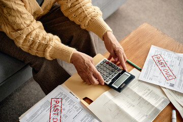 Senior woman using calculator while analyzing overdue invoices and bills at table, hands holding...