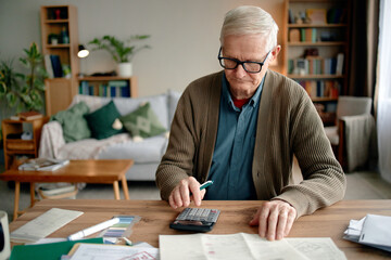 Senior man analyzing invoices and bills using calculator at desk, reviewing financial documents, sitting in home office environment, focused on paperwork