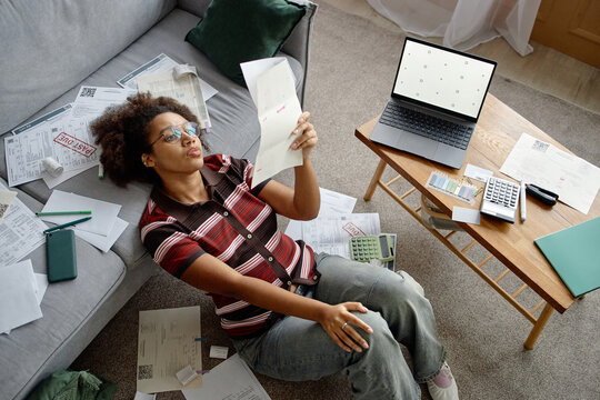 Young Black woman sitting on floor analyzing invoices and bills surrounded by paperwork using laptop and calculator financial documents scattered on table and couch