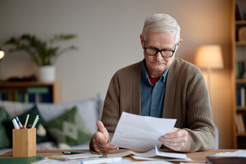 Senior man analyzing invoices and bills at desk, wearing eyeglasses, holding documents in hands, concentrating on paperwork, working in home office environment