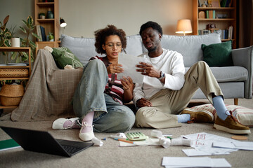 Young Black man and young woman sitting on floor analyzing invoices and bills together, using...