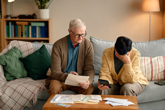 Senior man reading invoice while senior Asian woman holding calculator and covering face, both sitting on sofa analyzing bills and financial documents on coffee table