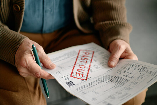 Senior man holding pen reviewing overdue invoice with past due stamp, analyzing financial document closely, focusing on payment details and outstanding bill
