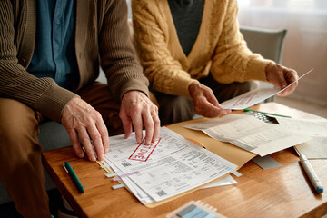 Senior man and senior woman reviewing overdue invoices and bills at table, sorting paperwork and...