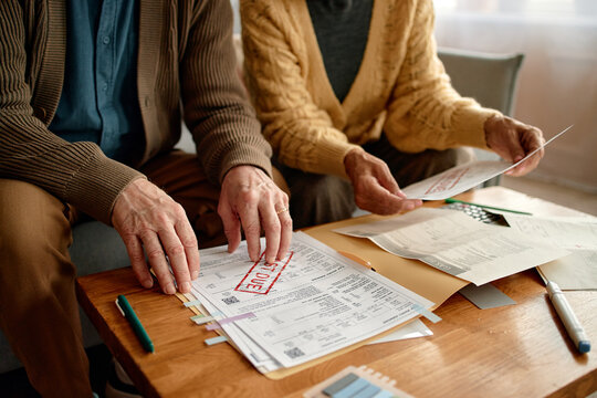 Senior man and senior woman reviewing overdue invoices and bills at table, sorting paperwork and using calculator, focusing on financial documents and household expenses