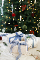 Wrapped stylish christmas presents with blue ribbons, green branches and scissors on festive paper against xmas tree with lights. Merry Christmas and Happy Holidays!