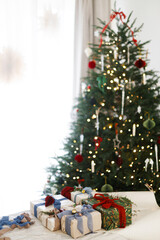  Merry Christmas and Happy Holidays! Wrapped stylish christmas presents with silk bows, green branch and berries, vintage ornaments on rustic table against decorated xmas tree with lights.