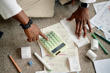 Black man analyzing invoices and bills using calculator, hands sorting financial documents on carpet, reviewing receipts and paperwork for expenses, pens and markers scattered around