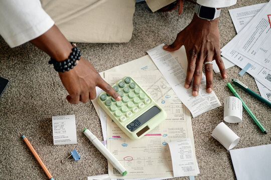 Black man analyzing invoices and bills using calculator, hands sorting financial documents on carpet, reviewing receipts and paperwork for expenses, pens and markers scattered around