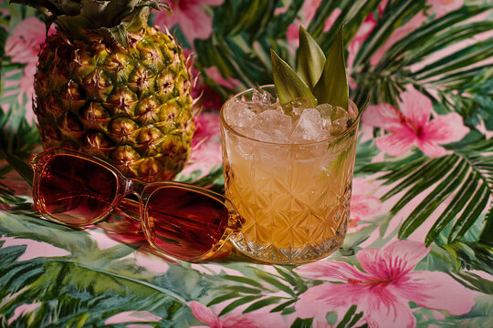 A glass of mixed drink with a pineapple and a pair of sunglasses on a table