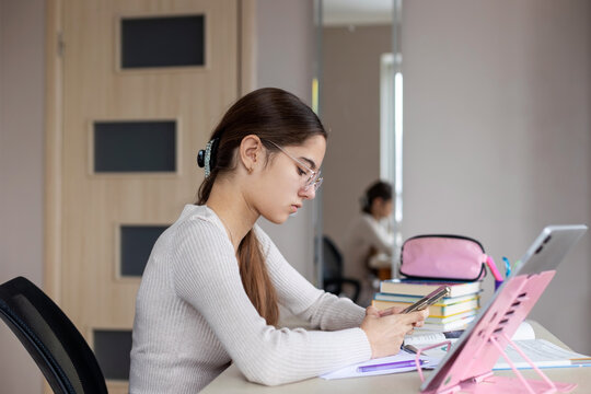 Teen girl studying with tablet and smartphone. Focused teenage girl uses smartphone while studying at desk with tablet, notebooks, and books during online or remote learning.
