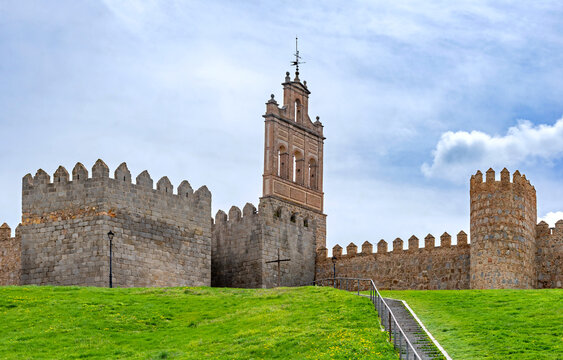 A view of the Walls of &Aacute;vila with stone towers lining a steep green hill, featuring a tall, slender reddish-brown stone chapel or watchtower built into the fortifications