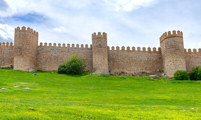 A portion of &Aacute;vila&rsquo;s ancient stone walls, with tall rounded towers rising above a bright green embankment and set beneath a softly clouded sky that creates a calm atmosphere