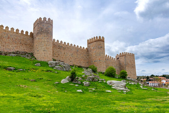 A portion of &Aacute;vila&rsquo;s ancient stone walls, with tall rounded towers rising above a bright green embankment and set beneath a softly clouded sky that creates a calm atmosphere