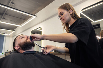 Woman Barber shaving bearded customer in modern style barbershop. Male Beard hair cutting with scissors. Man client visit gentleman grooming beauty salon for evens the beard