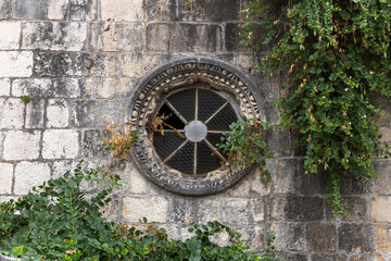 An ancient stone wall features a round, iron-grated window framed by carved stone