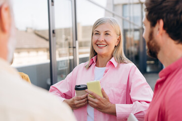 Cheerful gray haired woman holding coffee cup talking with bearded man outdoor on the street