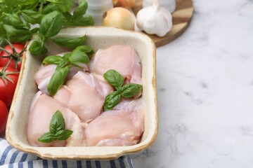 Raw chicken thighs with basil in baking dish and products on white marble table, closeup. Space for text
