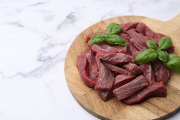 Pieces of raw beef meat and basil on white marble table, closeup. Space for text