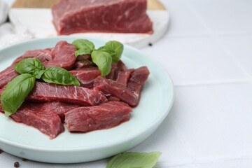 Pieces of raw beef meat and basil on white table, closeup. Space for text