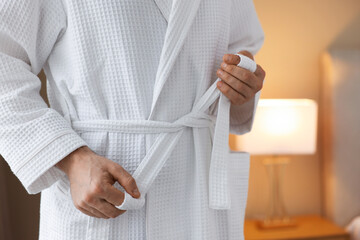 Man tying belt of bathrobe indoors, closeup