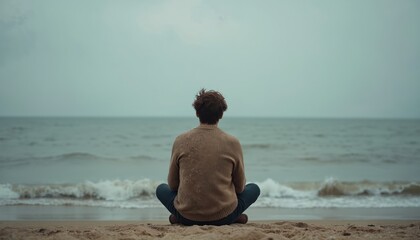 Lonely person sits on sandy beach. Ocean waves roll to shore under grey sky. Man looks out at sea. Contemplation and solitude fill scene. Peaceful, pensive feeling.