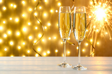 Merry Christmas and Happy New Year. Flute glasses with sparkling wine on white wooden table against golden background with festive decor and blurred lights, closeup. Space for text