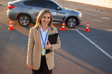 Fototapeta premium Smiling driving instructor holding clipboard near learner car on test site
