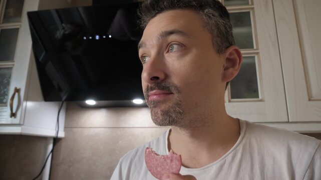 Man tastes salami sausage at home, enjoying the snack in a cozy kitchen with soft lighting