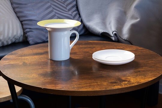 White ceramic mug with yellow rim and small plate on a rustic wooden table, surrounded by soft cushions, creating a cozy and inviting atmosphere for relaxation and enjoyment - Powered by Adobe