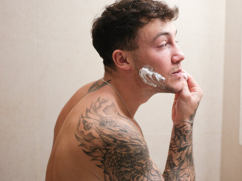 Young man with tattooed arms shaving in the bathroom. He applies shaving foam to his face and uses a razor while looking into the mirror, capturing a personal grooming and self-care moment - Powered by Adobe