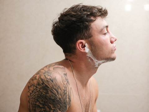 Young man with tattooed arms shaving in the bathroom. He applies shaving foam to his face while looking into the mirror, capturing a personal grooming and self-care moment.