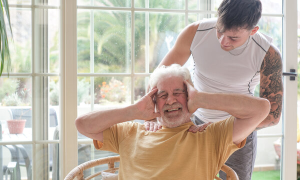 Young man massages the shoulders of an older man seated in a chair, creating a warm scene of caregiving and intergenerational support. Family connection, and well-being in a bright home environment.