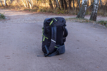 Black backpack with green accents is positioned on a dirt path surrounded by trees, showcasing outdoor adventure gear in a natural setting with autumn foliage