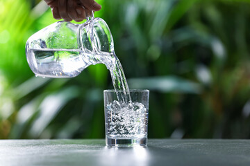 Woman pouring water into glass from jug at grey table against blurred green background, closeup. Space for text