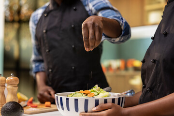 Young african american couple preparing food together in their kitchen, adding a pinch of salt to a fresh summer salad bowl. Man and woman enjoying a healthy and organic culinary activity.