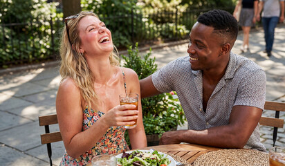 Cheerful young multiracial couple, a Black man and a blonde White woman, laughing heartily during a lunch date at an outdoor cafe table on a sunny summer day.