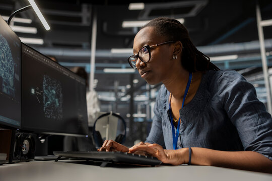 Focused data center developer using artificial intelligence deep learning tech. African american woman in server hub looking at AI programming language on PC monitor to update gear - Powered by Adobe