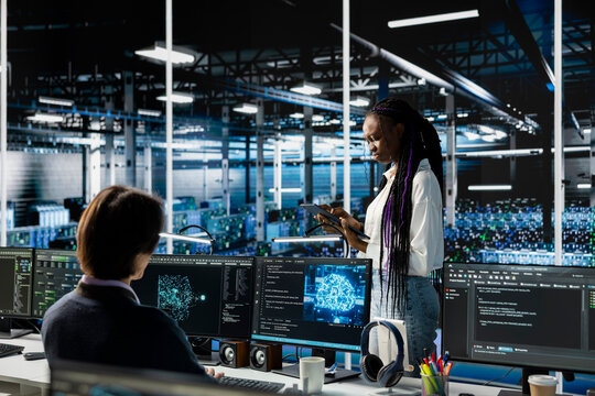System administrators in data center troubleshoot server connectivity issues using AI. African american woman and technician in server room auditing systems for vulnerabilities - Powered by Adobe