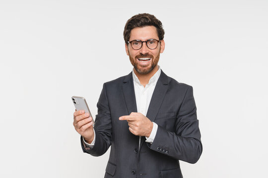 Business man wearing suit pointing on phone cellphone standing isolated on white background. Busy young businessman with smartphone in hands looking at camera, advertising mobile services, copy space