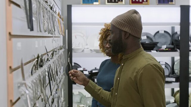 Medium side shot of smiling young African American couple standing by wall display at kitchenware shop, choosing cutlery, man holding up and inspecting knives and forks while discussing with wife